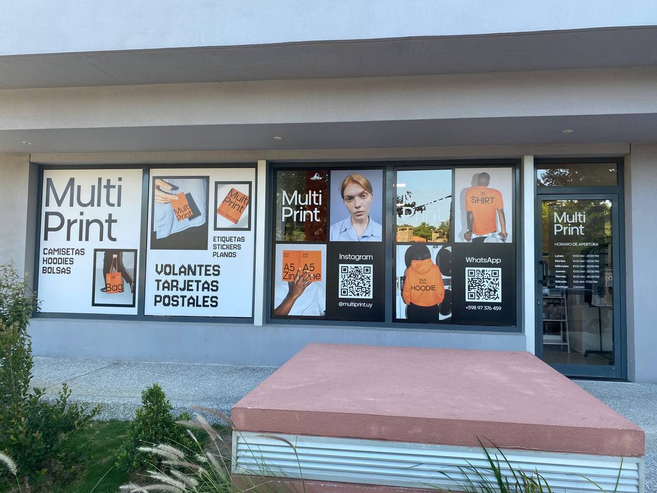 The MultiPrint shop window: posters of shirts, hoodies, bags and print materials, QR codes for Instagram and WhatsApp, the opening hours on the door